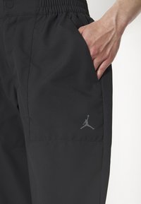 Pantalones deportivos negros con cintura elástica, bolsillos laterales y un logo gris de Jordan en el muslo izquierdo. Tela suave y ligera.