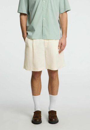 Pantaloni corti beige chiaro con pieghe frontali, abbinati a una camicia a maniche corte a righe verde e bianco, calze bianche e scarpe mocassino marroni.
