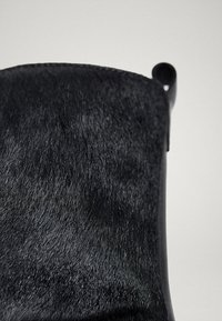 Gros plan sur la tige d'une botte en cuir noir texturé avec un bord supérieur roulé sur un fond gris clair uni.