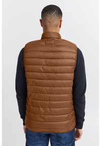 Braune Steppweste mit hohem Kragen, gestepptem Muster und schlankem Design. Getragen über einem dunklen Pullover, der einen sauberen Abschluss am Saum hat.