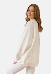 Biała, oversize bluza z opadłymi ramionami, miękką teksturą i ściągaczami. Noszona na białych legginsach, prezentująca luźny krój.