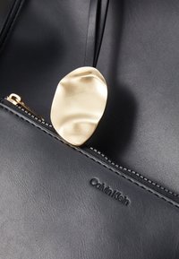 Čierna kožená taška s textúrovaným povrchom, so zlatým abstraktným ťaháčikom zipsu a embosovaným logom "Calvin Klein" na boku.