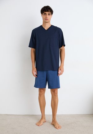 Dunkelblaues V-Ausschnitt-Shirt mit einer Brusttasche, kombiniert mit blauen gemusterten Shorts, die ein geometrisches Design aufweisen. Das Modell steht barfuß auf einem Teppich.