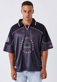 Camicia corta viola scuro con accenti rosa, colletto nero e un design grafico centrale circolare, realizzata in un tessuto liscio e lucido.