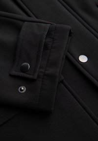 Tela negra con una textura suave; presenta un puño con botones y broches plateados y negros; líneas limpias y diseño ajustado.