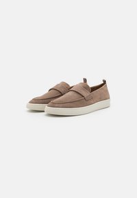 Chaussures slip-on en daim marron avec une finition lisse, dotées d'une large sangle et d'une semelle en caoutchouc blanche contrastante. Design minimaliste.