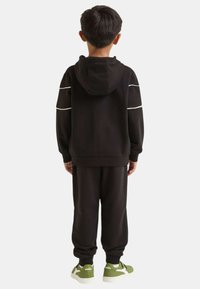 Felpa con cappuccio nera con strisce bianche, pantaloni neri abbinati e sneakers verdi e bianche. Il tessuto sembra morbido con una vestibilità comoda.