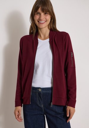 Femme souriante portant une veste zippée bordeaux sur une chemise blanche et un jean bleu foncé, debout devant un fond clair uni.