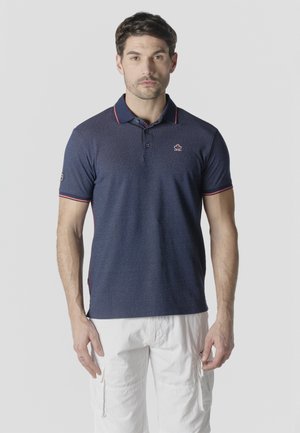 Egy férfi áll, sötétkék galléros pólót visel piros szegéllyel és fehér cargo nadrágot egy egyszerű, világos háttér előtt.