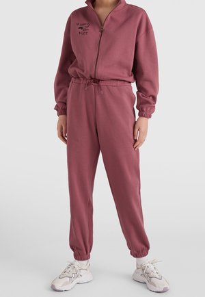 Osoba ubrana w mauve bluza rozpinana na zamek i dopasowane spodnie dresowe z napisem „Women of the Wave”, stojąca z rękami opartymi przy bokach, w białych trampkach.