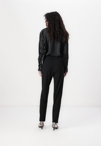 Zwarte satijnen blouse met lange mouwen en een ontspannen pasvorm, gecombineerd met strakke zwarte broek en zilveren hoge hakken, gezien vanaf de achterkant.