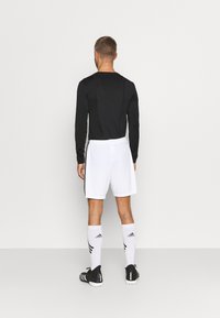 Maglietta nera a maniche lunghe, pantaloni corti bianchi con cinturino, calze da calcio bianche e nere, scarpe nere. Design atletico semplice, con accenti minimali.