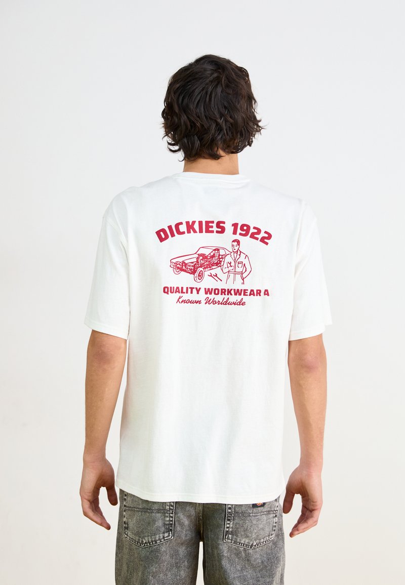 Valge puuvillane T-särk, mille seljaosal on punane graafiline tekst ja illustreerimine, millel on kirjas "DICKIES 1922 QUALITY WORKWEAR", lihtsa disainiga.