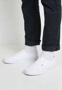 Weiße Leinenschuhe mit niedrigem Design, runder Zehenpartie und weißer Gummisohle. Getragen mit weißen Socken und dunklen Hosen, was einen lässigen Look zeigt.