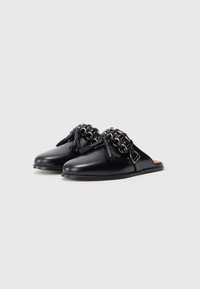 Mule-uri negre din piele cu slip-on, cu un accent dintr-un lanț texturat, ciucuri și o formă ușor curbată. Finisaj neted și talpă plată.