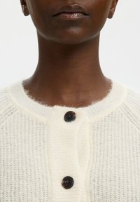 Nahaufnahme einer Person, die eine weiße Strickjacke mit zwei sichtbaren dunklen Knöpfen und einem abgerundeten Ausschnitt trägt.