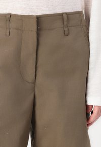 Brown cotton trousers with a smooth texture, featuring a waistband and side seams, partially shown with a hand near the pocket area.