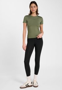 Olivgrünes, kurzärmeliges, figurbetontes Oberteil mit rundem Ausschnitt. Kombiniert mit hoch taillierten schwarzen Leggings und beigen Sportschuhen mit schwarzen Akzenten.