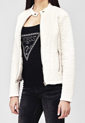 Veste matelassée blanche avec un col arrondi, fermeture éclair et motif en relief, portée sur un débardeur noir arborant un logo triangulaire.