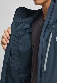 Blå vandtæt jakke indvendig med glat tekstur, lynlås-lommer og blødt for, med en lys grå lynlås og en mørk skjorte under.