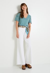 Top de color teal con escote en forma de V y mangas de ojales, combinado con jeans anchos blancos y un cinturón de bufanda estampada. Sandalias de plataforma marrones completan el look.