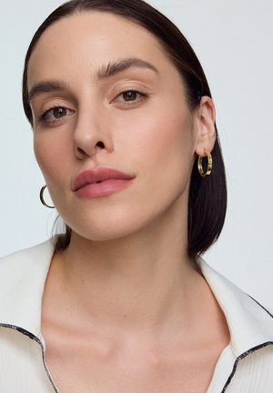 Złote kolczyki w kształcie kół, gładka jasna skóra i naturalny makijaż. Lekka tkanina z kołnierzykiem, charakteryzująca się miękkimi fakturami i subtelnymi kontrastami kolorów.