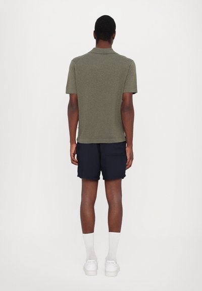 Camisa polo verde oliva com colarinho, combinada com calções azul-marinho. Meias brancas e sapatilhas completam o look. Vista traseira.
