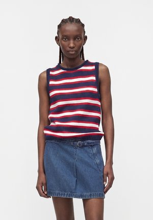 Młoda kobieta z warkoczami, ubrana w bezrękawnik w czerwono-biało-niebieskie paski i niebieską spódnicę jeansową z paskiem.