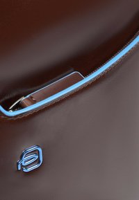 Borsa in pelle marrone con una texture liscia, caratterizzata da rifiniture blu e un logo nell'angolo inferiore. Include un dettaglio di una tasca con cerniera visibile.
