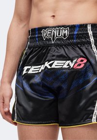 Άτομο που φοράει μαύρα σορτς Venum με λογότυπο Tekken 8, μπλε γραφικό σχέδιο και λευκές ρίγες με το όνομα της μάρκας στα πλαϊνά.