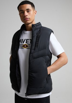Jonge man draagt een zwart gewatteerd vest over een wit T-shirt met het merk Zavetti Canada tegen een effen grijze achtergrond.