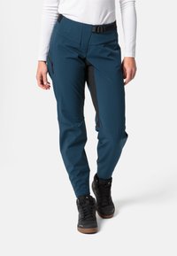 Teal buitenbroek met zwarte accenten, waterdichte stof, taps toelopende pasvorm en ritszakken. Draag het met zwarte wandelschoenen.