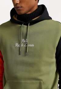 Bluza z kapturem w oliwkowej zieleni, z czarnymi i pomarańczowymi akcentami. Ozdobiona białym haftowanym logo oraz posiada przednią kieszeń. Tkanina o fakturowanej strukturze.