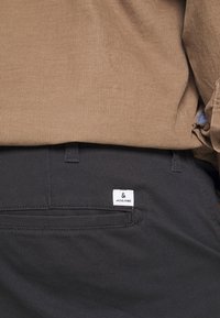 Pantalones negros con una textura suave, que cuentan con un bolsillo trasero y una pequeña etiqueta blanca que dice "JACK & JONES". La camisa es de canalé y de color marrón.