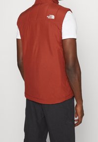 Červená bezrukávová vesta vyrobená z lehkého materiálu, s vysokým límcem a bílým logem na horní části zad. Hladká textura.