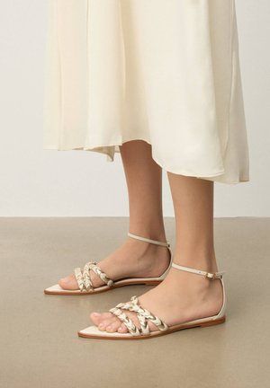 Fødder iført cremefarvede flade sandaler med flettede remme og ankelspænder, kombineret med en flydende elfenbensfarvet midikjole.