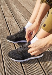 Personne portant des chaussettes blanches et des baskets noires en train de serrer ses lacets en étant assise sur une terrasse en bois en plein air.