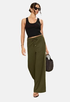 Mujer con camiseta sin mangas negra, pantalones anchos de pierna en verde oliva con cordón, gafas de sol negras, llevando un bolso tejido oscuro y sandalias negras.