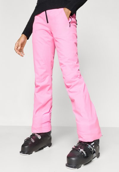 Roza smučarska hlače z gladko zunanjostjo, z zadrgami na žepih in ravnim krojem nog, združene s črnimi smučarskimi čevlji.