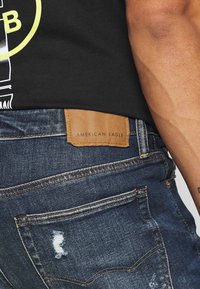 Mörkblå denimjeans med en sliten ficka, med en tan läderetikett präglad med "AMERICAN EAGLE." Beskuren vy som visar sidan av jeansen.