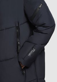 Navy puffer jacket with a quilted design, featuring a zippered pocket on the arm and contrasting black zippers and hardware.