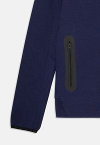 Vêtement à manches longues bleu marine avec des coutures visibles et une poche zippée noire verticale sur le côté près de l'ourlet.