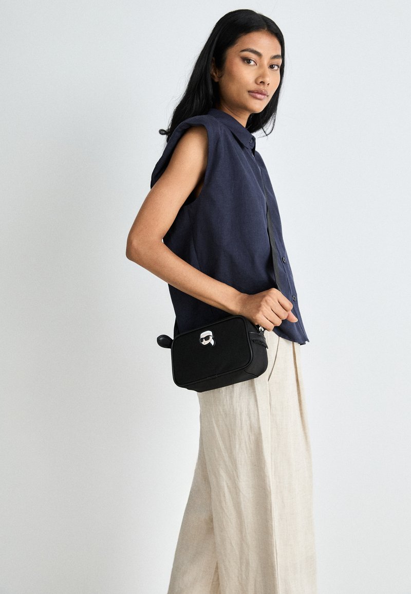 Borsa a tracolla nera con design di faccia da cartone animato, texture liscia e chiusura con zip, indossata con un top senza maniche blu scuro e pantaloni in lino chiaro.