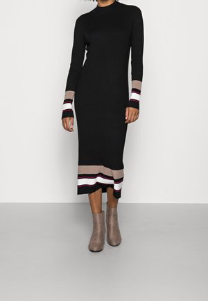 Kobieta w czarnej, prążkowanej dzianinowej sukience midi z beżowymi, białymi i bordowymi paskami na mankietach i dolnym brzegu, w połączeniu z taupe botkami za kostkę.