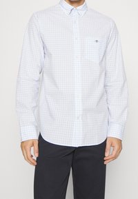 Světle modrá a bílá kostkovaná košile s dlouhými rukávy, s límcem a náprsní kapsou, vyrobená z prodyšné látky.