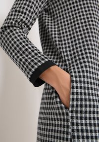 Ręka umieszczona w kieszeni czarno-białej koszuli z długim rękawem z detalem czarnego mankietu.