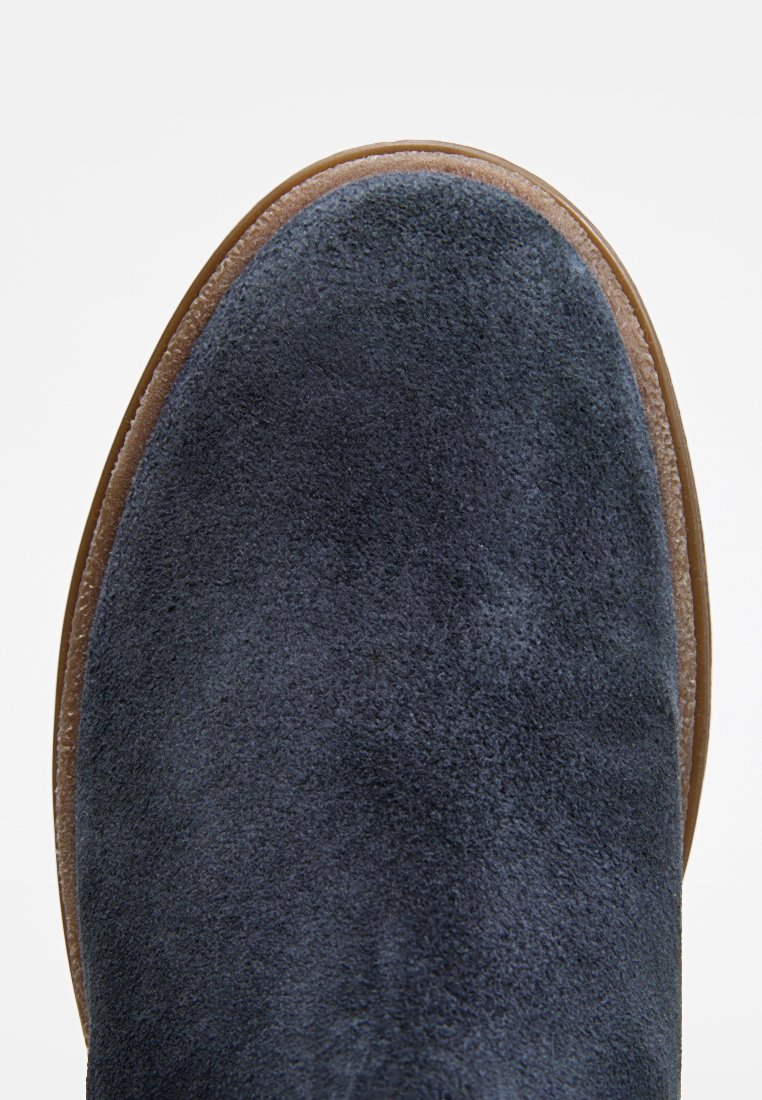 Marineblauer Wildlederstiefel mit runder Spitze und weicher Textur, ausgestattet mit einer beigefarbenen Gummisohle und gesteppten Details entlang der Kante.