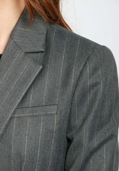 Blazer grigio a righe con revers a punta e tasca sul petto a soffietto indossato da una persona con lunghi capelli castani su uno sfondo neutro.