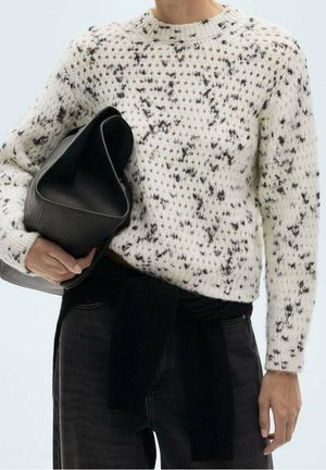 Person iført en hvid og sortspættet strikket sweater, der holder en stor sort lædertaske, med sorte bukser og et sort tøjstykke bundet om taljen.