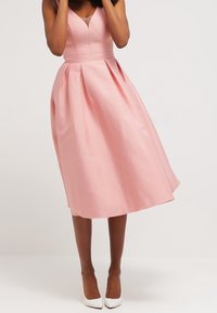 Vaaleanpunainen hihaton mekko, jossa on tiukka vartalo-osa, levenevä helma ja laskokset. Valmistettu sileästä kankaasta, siinä on V-aukko ja muotoiltu vyötärö.